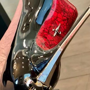 May include: A high-heeled shoe featuring a glossy black patent finish on the toe and heel, contrasted by a vibrant red panel with a black lace overlay. A silver-toned metallic accent is positioned on the side and at the base of the heel.