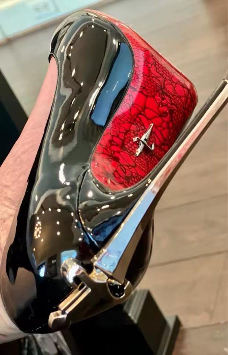 May include: A high-heeled shoe featuring a glossy black patent finish on the toe and heel, contrasted by a vibrant red panel with a black lace overlay. A silver-toned metallic accent is positioned on the side and at the base of the heel.