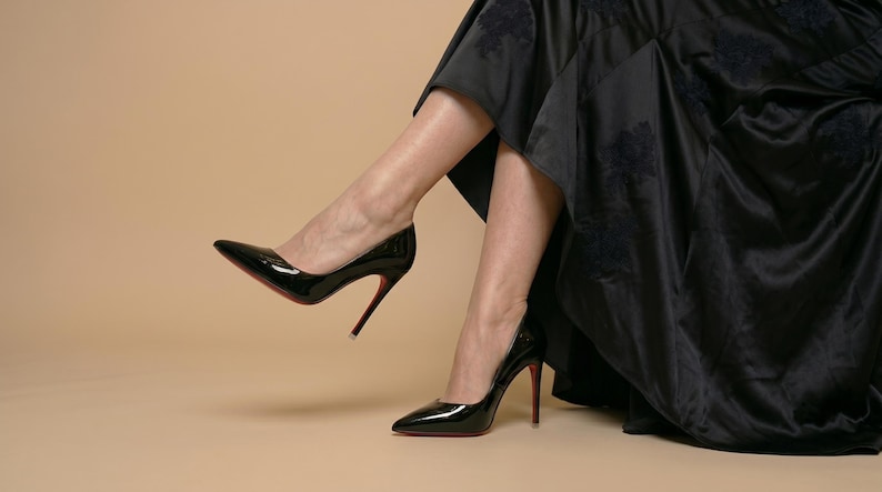 Puede incluir: Un par de zapatos de tac&oacute;n de aguja en cuero negro brillante con punta afilada y suelas rojas distintivas, combinados con una falda larga y fluida de sat&eacute;n negro con un sutil patr&oacute;n floral tonal en la tela.