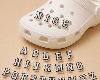 May include: A white foam clog shoe featuring circular ventilation holes, decorated with the word NICE in black and white varsity-style letter charms. Below the shoe are individual alphabet letter charms from A to Z in the same style.