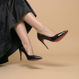Puede incluir: Un par de zapatos de tac&oacute;n alto de charol negro con punta afilada y tacones de aguja finos. Las suelas de los zapatos son de un color rojo vibrante. Los zapatos se llevan con una falda larga y fluida de color negro con un patr&oacute;n floral.