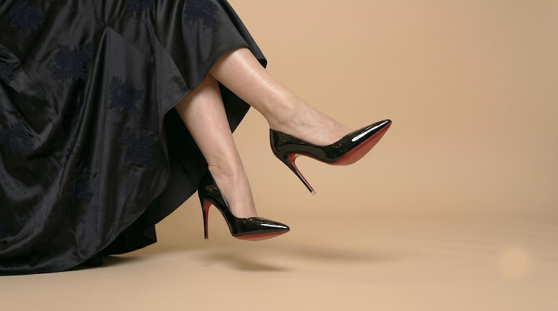 Puede incluir: Un par de zapatos de tac&oacute;n alto de charol negro con punta afilada y tacones de aguja finos. Las suelas de los zapatos son de un color rojo vibrante. Los zapatos se llevan con una falda larga y fluida de color negro con un patr&oacute;n floral.