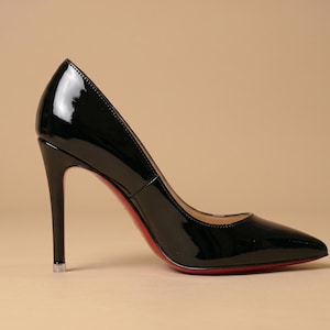 Puede incluir: Un zapato de tac&oacute;n de charol negro con punta afilada y tac&oacute;n de aguja alto. El zapato presenta un acabado brillante y reflectante, con una suela roja distintiva visible a lo largo del borde inferior, mostrando una silueta elegante y definida.