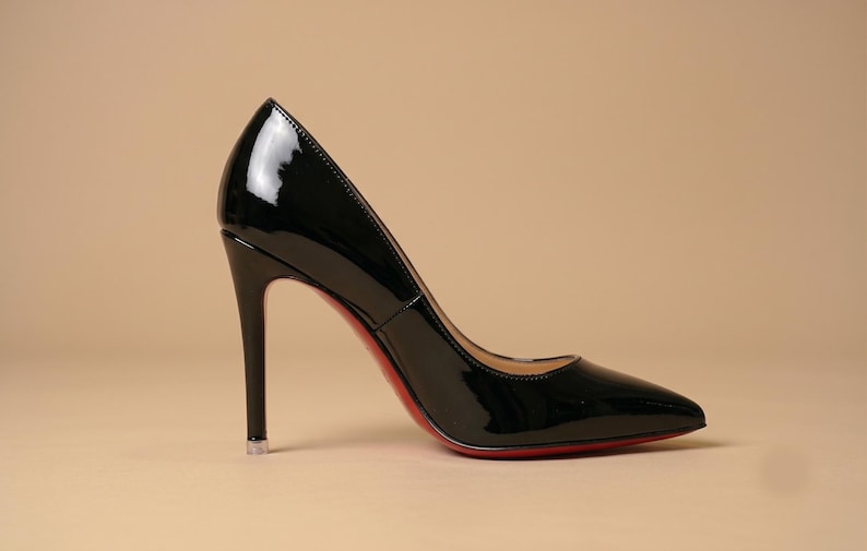 Puede incluir: Un zapato de tac&oacute;n de charol negro con punta afilada y tac&oacute;n de aguja alto. El zapato presenta un acabado brillante y reflectante, con una suela roja distintiva visible a lo largo del borde inferior, mostrando una silueta elegante y definida.