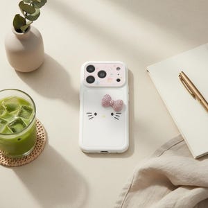May include: A white smartphone case featuring a stylised cat face with black whiskers and a small yellow nose. The case includes a three-dimensional pink textured bow attached to the back and a light pink grid pattern surrounding the camera lens area.