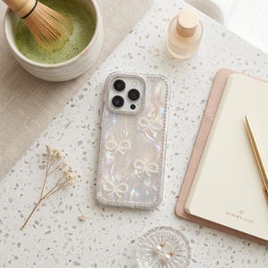 May include: A pearlescent phone case with bow accents and a crystal border. The case is next to a pink notebook, a gold pen, and a small glass dish with earrings. A bowl of matcha tea and a small bottle are also in the image.
