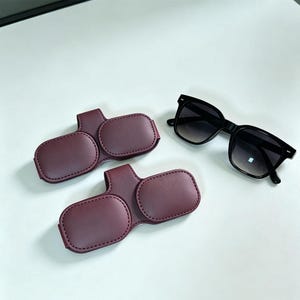 May include: Two maroon faux leather magnetic eyeglass holders with a smooth texture and stitched edges, positioned next to a pair of black-framed sunglasses with dark gradient lenses. The holders feature a rectangular shape with a central tab.