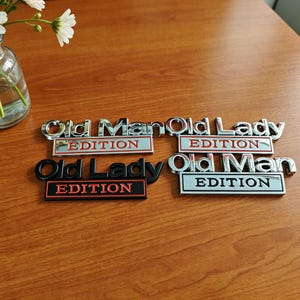 Puede incluir: Cuatro emblemas metálicos rectangulares con reverso adhesivo. Dos emblemas cromados dicen Old Man Edition y Old Lady Edition con texto en rojo. Un emblema negro dice Old Lady Edition con texto en rojo. Un emblema cromado dice Old Man Edition con texto en negro.