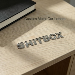 Puede incluir: Letras de coche metálicas brillantes que deletrean "SHITBOX" dispuestas sobre una superficie de madera clara. El texto "Custom Metal Car Letters" está impreso encima de las letras. Un libro negro es visible en el fondo.