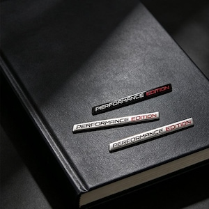 Puede incluir: Tres emblemas adhesivos rectangulares con el texto PERFORMANCE EDITION en una fuente sans-serif moderna. Un emblema es negro con letras blancas y rojas, y dos son plateados con letras negras y rojas, dispuestos sobre un cuaderno de color negro.