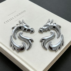 May include: Two metallic silver dragon emblems with a polished finish. Each dragon is depicted in a stylised, curved posture with sharp, angular features, including pointed horns and a sweeping tail. The emblems are placed on a white book cover.