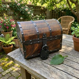 May include: A dark brown wooden chest with a rounded lid, featuring decorative black metal bands with embossed patterns, metal studs, and a large black metal padlock attached to the front latch mechanism, resting on a weathered wooden surface.