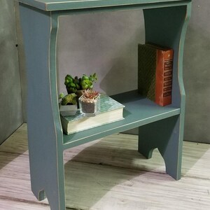 Pode incluir: Uma mesa lateral de madeira azul-esverdeada com um tampo retangular e uma prateleira inferior. A mesa apresenta bordas desgastadas e suportes laterais curvos. Na prateleira inferior, h&aacute; um livro com capa estampada, duas pequenas suculentas em vasos e um livro laranja.