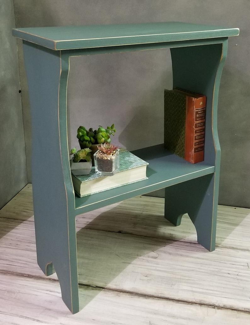 Pode incluir: Uma mesa lateral de madeira azul-esverdeada com um tampo retangular e uma prateleira inferior. A mesa apresenta bordas desgastadas e suportes laterais curvos. Na prateleira inferior, h&aacute; um livro com capa estampada, duas pequenas suculentas em vasos e um livro laranja.