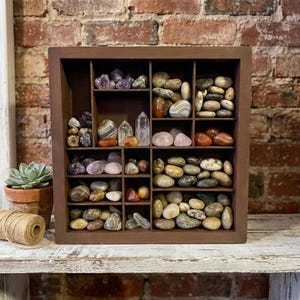 May include: A dark brown wooden shadow box display case with multiple square and rectangular compartments filled with an assortment of polished stones, crystals, and pebbles in various colors including white, gray, brown, orange, and purple.
