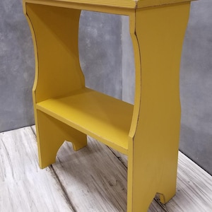 Pode incluir: Uma mesa lateral de madeira amarelo-mostarda com um tampo retangular, uma prateleira inferior &uacute;nica e suportes laterais curvos. As bordas da madeira pintada de amarelo mostram sinais de desgaste, revelando um tom mais escuro por baixo.