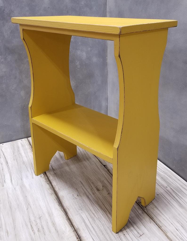 Pode incluir: Uma mesa lateral de madeira amarelo-mostarda com um tampo retangular, uma prateleira inferior &uacute;nica e suportes laterais curvos. As bordas da madeira pintada de amarelo mostram sinais de desgaste, revelando um tom mais escuro por baixo.