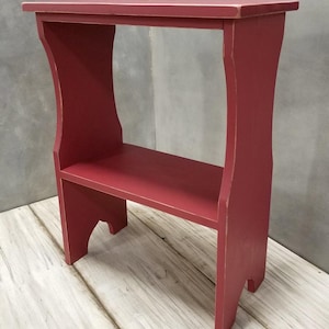 Pode incluir: Uma pequena mesa lateral retangular de madeira pintada com acabamento vermelho fosco. A pe&ccedil;a apresenta uma superf&iacute;cie superior plana, uma prateleira inferior &uacute;nica e dois pain&eacute;is laterais verticais com recortes arqueados na base, mostrando um leve desgaste.