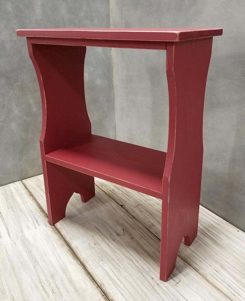Pode incluir: Uma pequena mesa lateral retangular de madeira pintada com acabamento vermelho fosco. A pe&ccedil;a apresenta uma superf&iacute;cie superior plana, uma prateleira inferior &uacute;nica e dois pain&eacute;is laterais verticais com recortes arqueados na base, mostrando um leve desgaste.