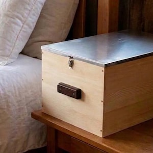 May include: A rectangular storage chest constructed from light-colored wood with a smooth, flat metal lid. The chest features a small metal latch on the front center and a dark brown horizontal handle mounted on the side panel for easy carrying.