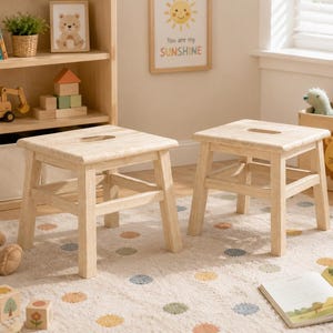 May include: Two light-colored wooden stools with four sturdy legs and a rectangular seat featuring a central cutout handle. The stools have a smooth, natural finish and are positioned on a light-colored rug with a pattern of small, multicolored dots.