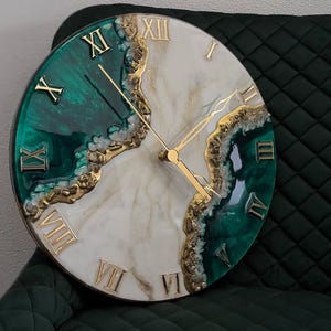 Può includere: Un orologio da parete rotondo con un design diviso in resina verde smeraldo intenso e bianco effetto marmo. La superficie è accentuata da dettagli in foglia oro testurizzata lungo la divisione diagonale, numeri romani e lancette dorate.