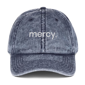 Könnte beinhalten: Eine dunkelblaue Baseballkappe in Acid-Wash-Optik mit sechs Paneelen und gebogenem Schirm. Das Wort mercy. ist in weißen Kleinbuchstaben mittig auf der Vorderseite eingestickt, ergänzt durch einen Knopf oben und gestickte Ösen.