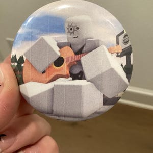 May include: A circular pin-back button featuring a graphic illustration of a blocky, white character with a sun-shaped eye symbol playing an orange acoustic guitar. The character is surrounded by large white cubes against a blue sky and green trees.