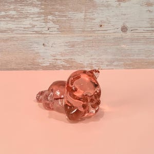 May include: A translucent pink glass figurine shaped like a small bear lying down in a resting position. The bear has smooth, rounded features and a glossy finish that reflects light, showcasing the clear, tinted material of the sculpture.