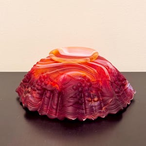 May include: A translucent red and orange glass bowl featuring a raised relief pattern of grape clusters and leaves. The bowl has a scalloped edge and a circular base, with a smooth, glossy finish that transitions from deep crimson to bright amber.