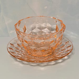 May include: A translucent peach-colored glass bowl and matching saucer set. Both pieces feature a repeating geometric diamond-cut pattern across their surfaces, creating a faceted texture that catches the light with a soft, warm glow.