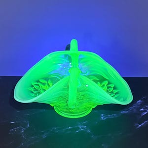 May include: A translucent glass basket glowing with a vibrant neon green hue under ultraviolet light. The basket features a central arched handle and a wide, scalloped rim with embossed floral patterns on the interior base and sides.