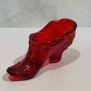 May include: A translucent ruby red glass shoe figurine featuring intricate embossed scrollwork and floral patterns across the exterior surface. The decorative object is shaped like a high-heeled boot with a pointed toe and a wide opening at the top.
