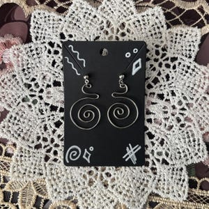 Peut inclure: Boucles d'oreilles argentées en forme de spirale sur une carte noire. Les boucles d'oreilles ont un fermoir à tige et un design en fil de fer formant une spirale. La carte est noire avec des gribouillis blancs.