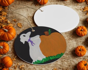 May include: A Halloween-themed ceramic ornament featuring a ghost and a pumpkin design. The ghost is white with a purple bow, and the pumpkin is orange. The ornament is round with a string for hanging. Surrounding the ornament are small pumpkins.