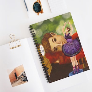 Puede incluir: Cuaderno de espiral con una ilustración colorida de una bailarina con tutú morado, un hombre y un reloj. El cuaderno tiene una encuadernación en espiral negra y está abierto sobre una superficie blanca con un clip dorado.
