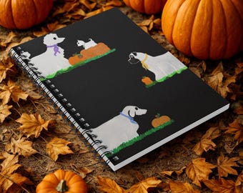 May include: A spiral-bound notebook with a black cover featuring illustrations of white dogs with pumpkins. The notebook is surrounded by orange pumpkins and autumn leaves, creating a fall theme. The dogs are wearing colorful accessories.