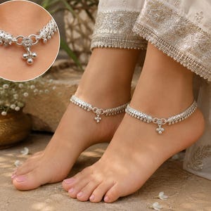 May include: A pair of silver anklets featuring a woven chain design with small dangling spherical charms along the edge and a central floral-shaped link with a three-bead pendant. The metallic finish is bright and reflective in the light.