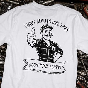 Puede incluir: Camiseta blanca con una ilustración en blanco y negro de un mecánico haciendo una señal de pulgar hacia arriba. El texto dice "I don't always lose tools, just the 10mm." La camiseta se muestra en una caja de herramientas llena de herramientas.