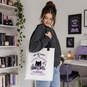 May include: A white tote bag featuring a black cat illustration sitting at a desk with books, a lamp, and a mug. The text reads: ASK ME ABOUT MY CURRENT OBSESSION BUT UNDERSTAND THIS IS A THREAT. The design uses purple and black accents.