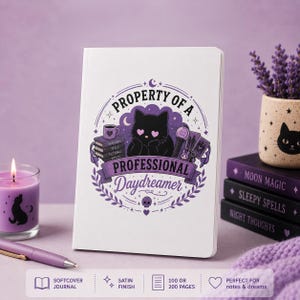 May include: A white softcover journal featuring a purple graphic of a black cat with heart-shaped eyes, surrounded by books, a mug, and a light stick. The text reads Property of a Professional Daydreamer. The journal has a satin finish.