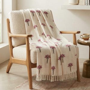 May include: A cream-colored throw blanket featuring a repeating pattern of small, dark pink lotus flower motifs. The blanket has a textured fabric finish and is finished with short, knotted fringe along the bottom edge, draped over a chair.