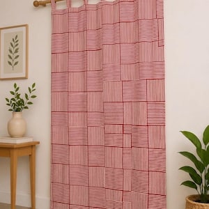 Op de afbeelding: Een rechthoekig gordijn met een geometrisch patchworkpatroon van rode en witte strepen. De stof hangt aan een houten roede door middel van een reeks stoffen lussen langs de bovenrand. De strepen in elk blok wisselen van richting.