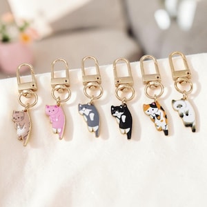Pode incluir: Uma coleção de seis chaveiros em forma de gato. Cada chaveiro apresenta um pingente de gato de cores diferentes, incluindo rosa, cinza, preto e tartaruga, pendurado em um fecho dourado. Os chaveiros são exibidos em uma superfície branca.