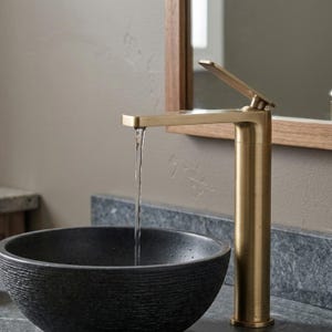 May include: A tall, brushed brass bathroom tap with a single lever handle, dispensing a steady stream of water into a dark, textured, circular stone vessel sink. The tap features a sleek, cylindrical body and a flat, horizontal spout.