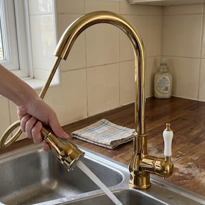 Könnte beinhalten: Ein polierter goldener Küchenwasserhahn mit hohem Auslauf und ausziehbarer Brause. Der Wasserhahn verfügt über einen weißen Keramikhebelgriff und einen flexiblen goldenen Schlauch, der einen stetigen Wasserstrahl in ein Edelstahlbecken abgibt.