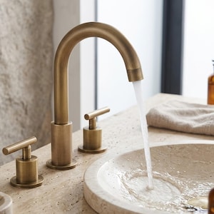Puede incluir: Un grifo de baño de latón cepillado con caño de arco alto y dos manijas separadas en forma de T. El agua fluye desde el caño hacia un lavabo de piedra circular. El grifo presenta un acabado metálico y un diseño de líneas cilíndricas.