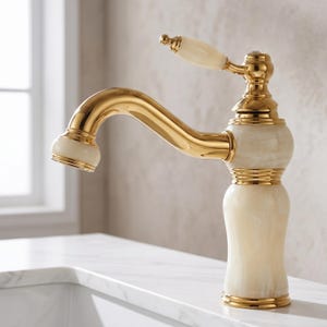 May include: A bathroom tap featuring a polished gold finish combined with cream-coloured stone accents. The design includes a curved spout, a single lever handle with a stone inlay, and a cylindrical base with a smooth, marbled stone texture.