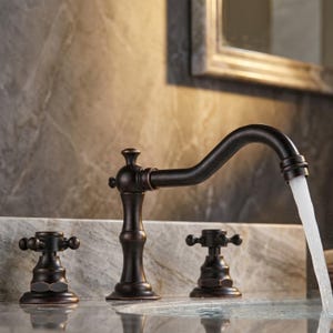 May include: A dark bronze bathroom tap with a curved spout and two cross-handle knobs. Water flows from the spout into a sink basin. The tap features a metallic finish with a smooth, curved design and distinct cross-shaped handles.