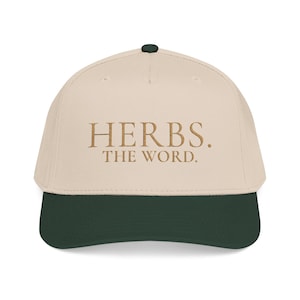 May include: A beige baseball cap with a dark green brim and a dark green button on top. The front of the cap features embroidered text in a brown serif font that reads HERBS. THE WORD. The cap has a structured crown and stitched eyelets.
